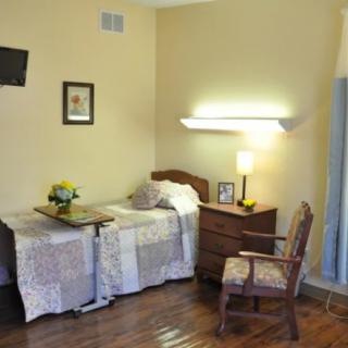 A cozy bedroom or hospital room with a bed, dresser, chair, and mounted TV.