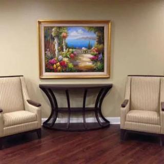 An elegant room interior featuring two beige upholstered chairs flanking a dark wood console table, topped with a colorful landscape painting, all set on a polished wooden floor.