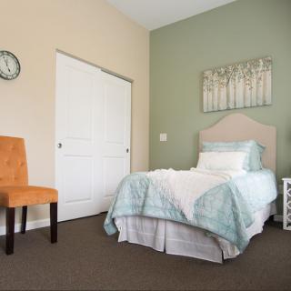 A cozy bedroom featuring a single bed with light blue bedding, an orange chair, and minimalist wall art.