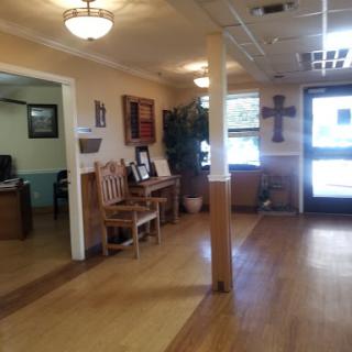 A cozy waiting area featuring wooden floors and chairs, with decor including plants and a cross.