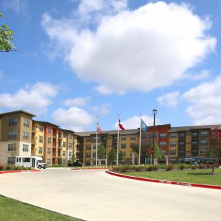 A modern residential building under a blue sky with scattered clouds, featuring multiple flags and a driveway.