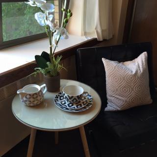 A cozy corner with a small table set for tea, accompanied by a potted plant and cushioned chair beside the window.
