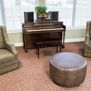 A cozy living room with a piano, two chairs, and a round ottoman.