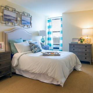 A cozy bedroom featuring a neatly made bed with decorative pillows, flanked by two nightstands and lamps. The room is softly lit and has a window with patterned curtains.