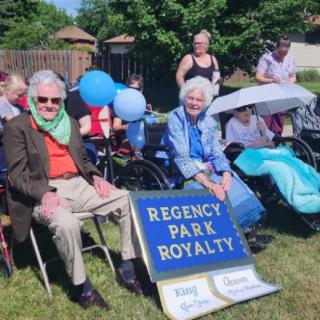 A group of elderly people sitting outdoors in wheelchairs participating in a community event titled 'Regency Park Royalty'.