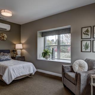 A modern bedroom with minimalist decor, featuring a cozy bed, armchair, and ample natural light from a large window.