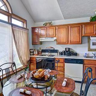 A cozy kitchen with wooden cabinets and a glass dining table set for a meal, illuminated by natural light from a large window.