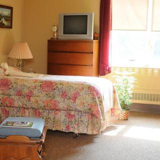 A cozy bedroom featuring a floral-patterned bed, dresser with a television on top, and a lamp by the window.
