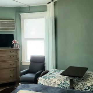 A cozy bedroom featuring a bed with floral bedding, a reclining chair, a wooden dresser with a TV on top, and a large window with curtains.