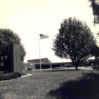 A black and white photo of a building entrance with a flag flying, surrounded by greenery.
