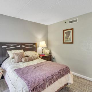 A cozy bedroom with a neatly made bed, featuring a purple throw blanket and multiple pillows. A lamp and decor sit on the nightstand.