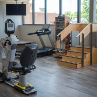 A rehabilitation center room equipped with exercise machines, including a treadmill, recumbent stepper, and therapy stairs.