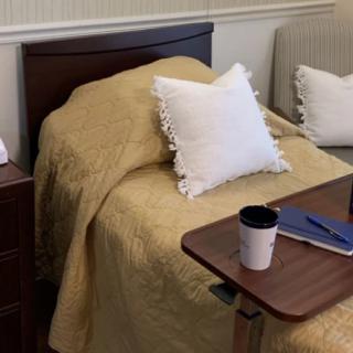 A hospital room setup with a bed, pillow, and side table holding a cup and notebook.