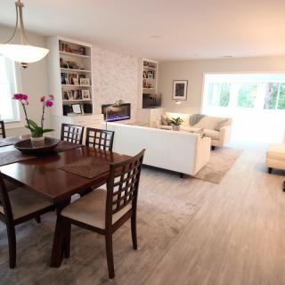 An open-concept living and dining area with modern interior design, featuring a wooden dining table, white sofas, and natural light.