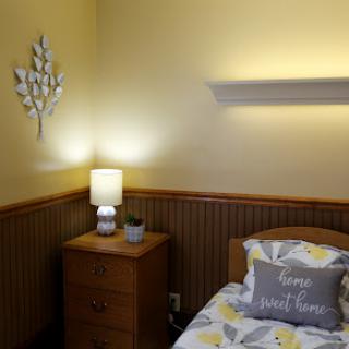 A cozy and well-decorated bedroom with yellow walls, featuring a nightstand with a lamp and decorative bedding.