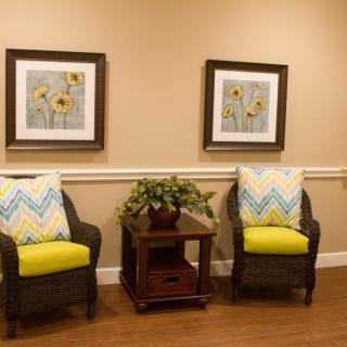 A cozy seating area with two wicker chairs, adorned with yellow and chevron pillows, flanking a wooden side table topped with a plant. The walls are decorated with floral artwork.