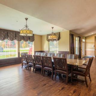 A spacious dining room with a large wooden table and chairs, illuminated by chandeliers and natural light through large windows.