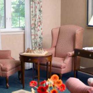 A cozy living room corner with armchairs, a chessboard on a side table, and a window with floral curtains.