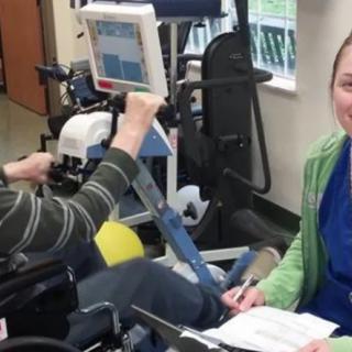 A person in a wheelchair using exercise equipment with assistance from a healthcare professional.