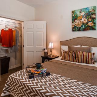 A cozy bedroom featuring a neatly made bed with striped pillows and patterned bedding, an open closet displaying clothes, and decorative elements like a painting and lamp.