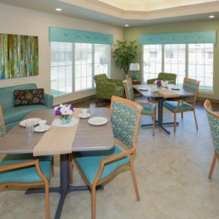 A cozy dining area with tables and chairs, featuring large windows and indoor plants.