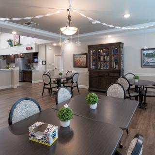 A cozy dining area with elegant decor, featuring multiple tables and chairs, a chandelier, and potted plants on wooden flooring.