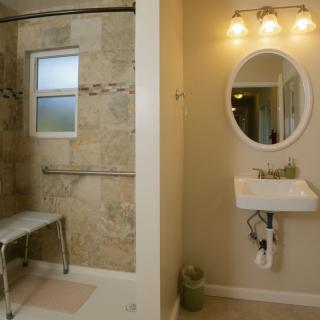 An accessible bathroom featuring a walk-in shower with a grab bar and a bench, alongside a wall-mounted sink and oval mirror under warm lighting.