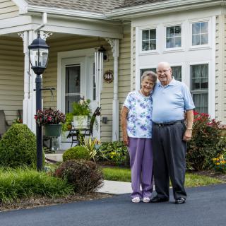 A smiling elderly couple standing together in front of their house.