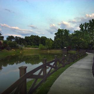 A peaceful park with a winding path alongside a calm body of water, bordered by trees and a wooden fence.