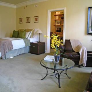 A cozy bedroom with a neatly made bed, glass coffee table, and comfortable chair, accented by warm yellow walls and decorative touches.
