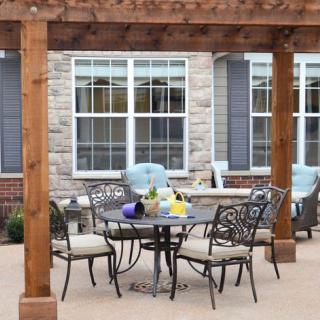 An inviting patio setup with a table and chairs under a wooden pergola against a brick house exterior.