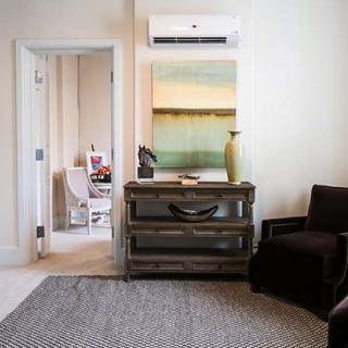 A cozy and stylish living room featuring a comfortable chair, industrial-style dresser, and modern art on the wall beneath an air conditioning unit.