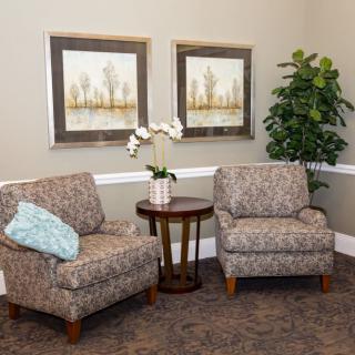 A cozy living room corner with two patterned armchairs, a small round table with a white orchid, framed artwork on the wall, and a potted plant.