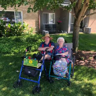 Two elderly individuals sitting outside in wheelchairs, enjoying a sunny day in the garden.