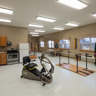 A well-lit exercise room featuring a recumbent bike, rehabilitation bars, and a small kitchen area.