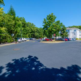 A sunny parking lot with cars, surrounded by trees and a white building.