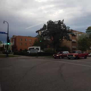 A view of an urban intersection with buildings and parked vehicles under a cloudy sky.