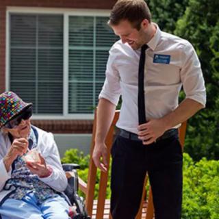A caregiver interacting with an elderly person seated in a wheelchair outdoors.