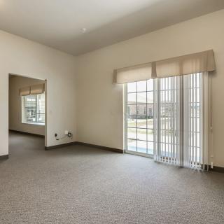 A spacious, empty living room with large windows and neutral carpeting, allowing ample natural light.