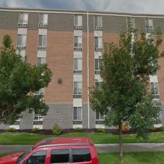 A multi-story apartment building with a brick facade, surrounded by trees and a red car parked in front.