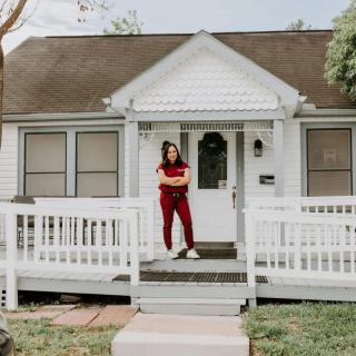 A person stands on the porch of a small white house.