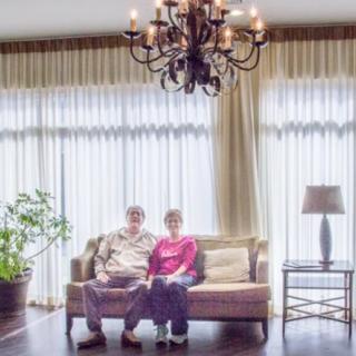 An elderly couple sitting on a sofa in a bright living room with large windows and elegant decor.