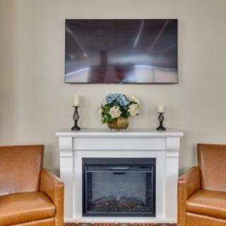 A cozy living room setup featuring a white fireplace, two leather armchairs, a television mounted above the mantel, and decorative elements including flowers and candles.