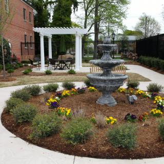 A neatly landscaped garden featuring a central fountain and a pergola in the background, surrounded by vibrant flowers and pathways.