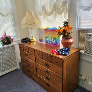 A cozy interior scene featuring a wooden dresser with decorative items, including a lamp, vase of flowers, and a colorful gift bag.