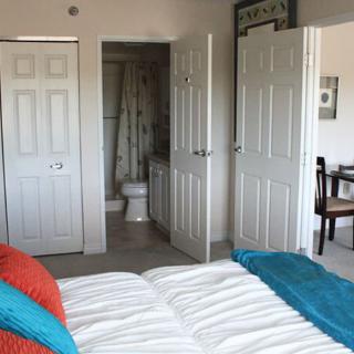 A cozy bedroom with colorful pillows, open doors leading to a bathroom, and a glimpse of a small desk area.