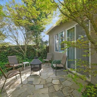 A cozy patio area with seating around a fire pit, surrounded by lush greenery and part of a house exterior.