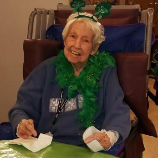 An elderly woman dressed in festive green accessories smiles while sitting indoors, possibly celebrating St. Patrick's Day.