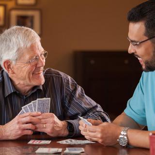 Two men playing cards and smiling at each other indoors.