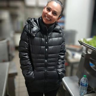 A cheerful woman wearing a black puffer jacket stands in a kitchen, radiating positivity.